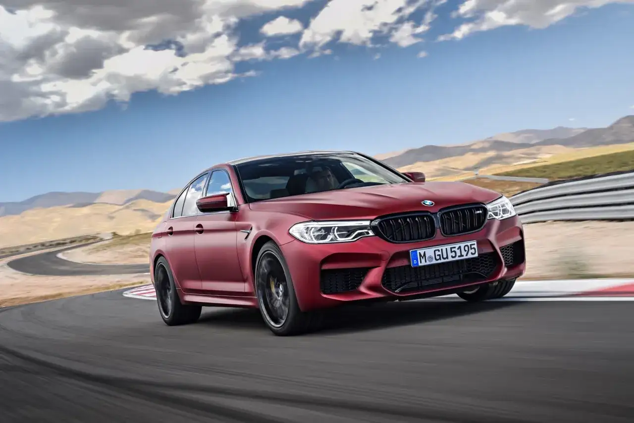 Czerwone BMW M5 Competition na torze wyścigowym, z widocznymi śladami opon na asfalcie i górzystym krajobrazem w tle.