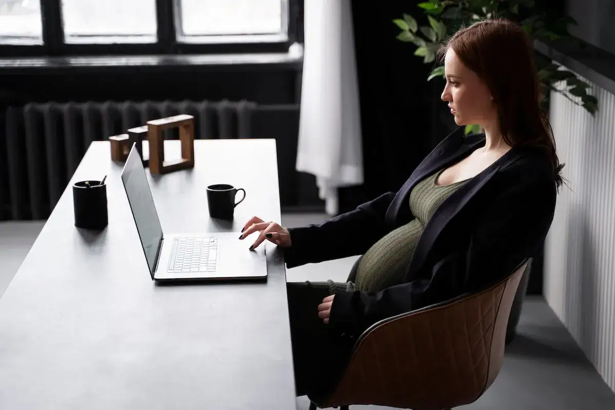 Kobieta w l4 w ciąży pracuje przy laptopie. Na biurku kubek i ozdoby.