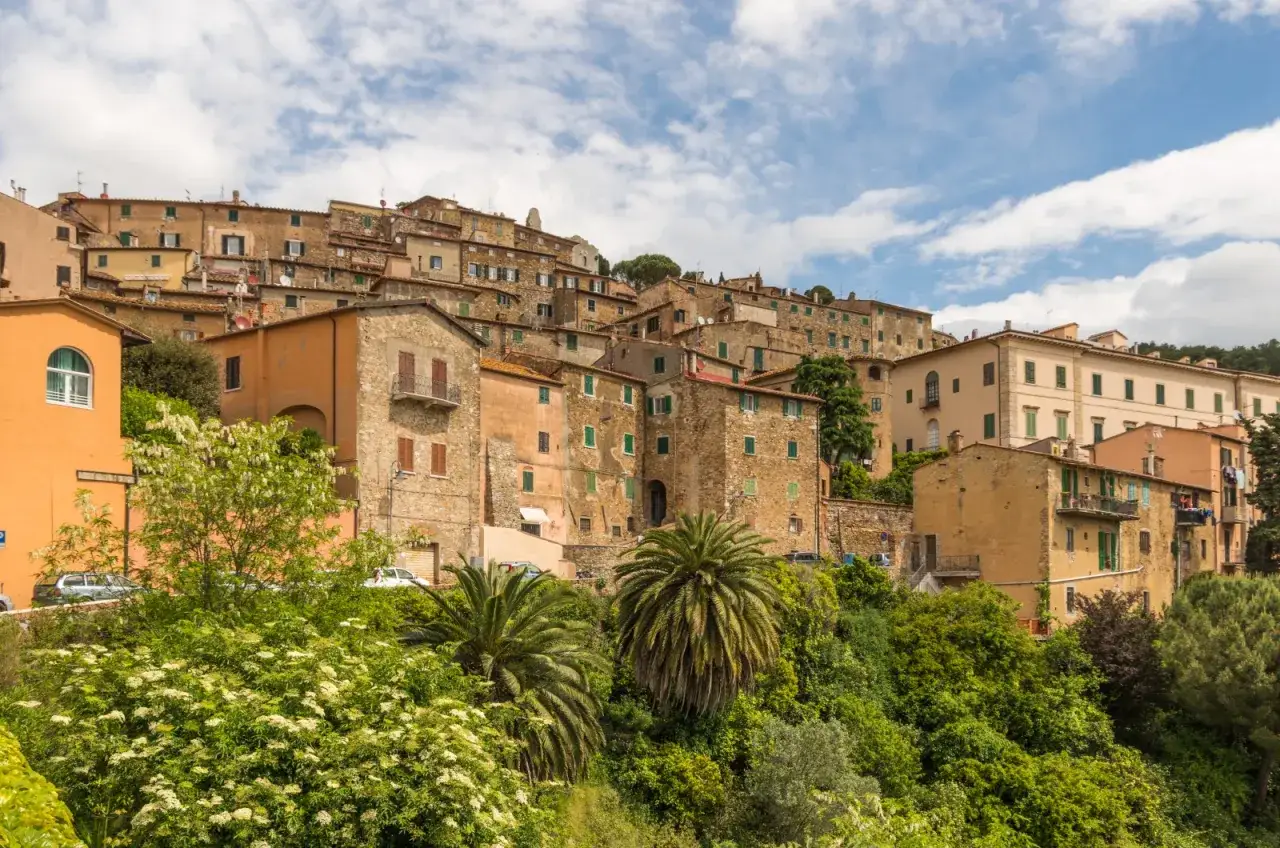 Case colorate e pietra si arrampicano sulla collina di Campiglia Marittima, tra palme e vegetazione rigogliosa.