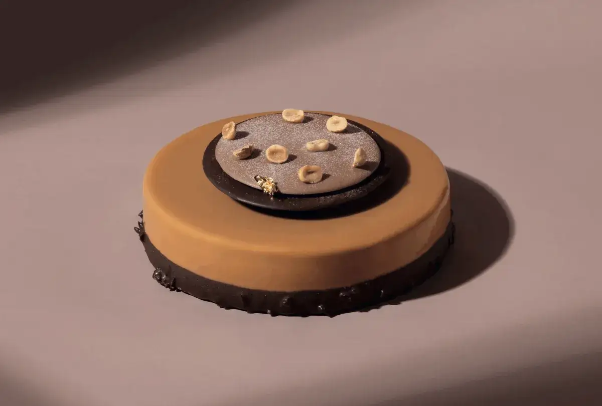 Torta lucida color caramello con decorazione centrale di nocciole e foglia d'oro, che evoca la ricchezza della pasta di nocciole.