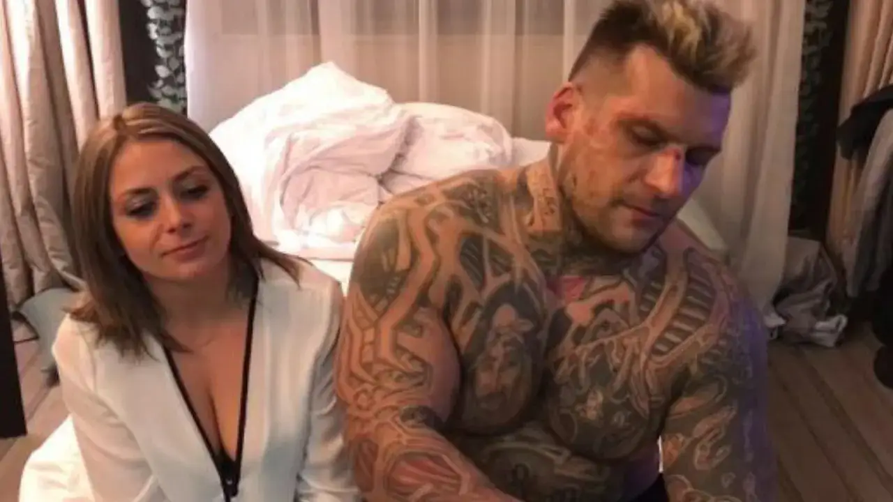 Popek z żoną siedzą na łóżku. Ona ma na sobie białą bluzkę, on jest bez koszulki, pokryty tatuażami.