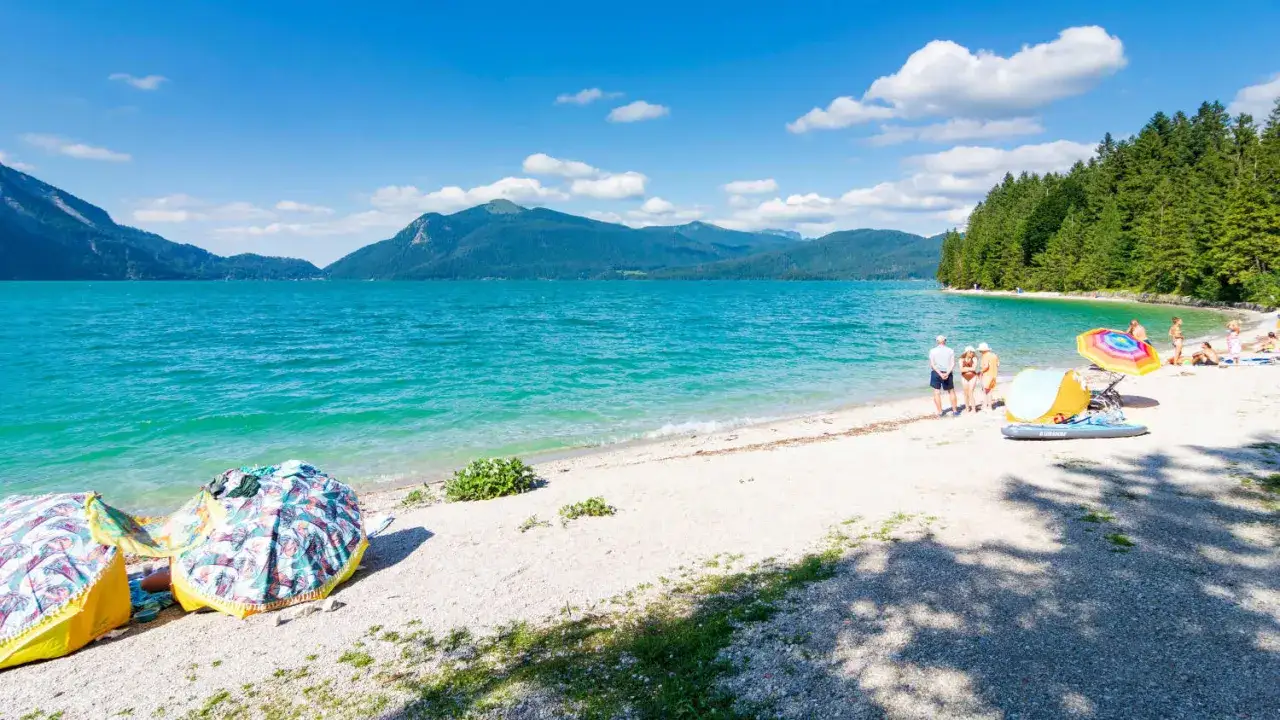 Ein sonniger Tag am badesee bayern. T&uuml;rkises Wasser, Berge und ein Strand mit Menschen, die sich entspannen.