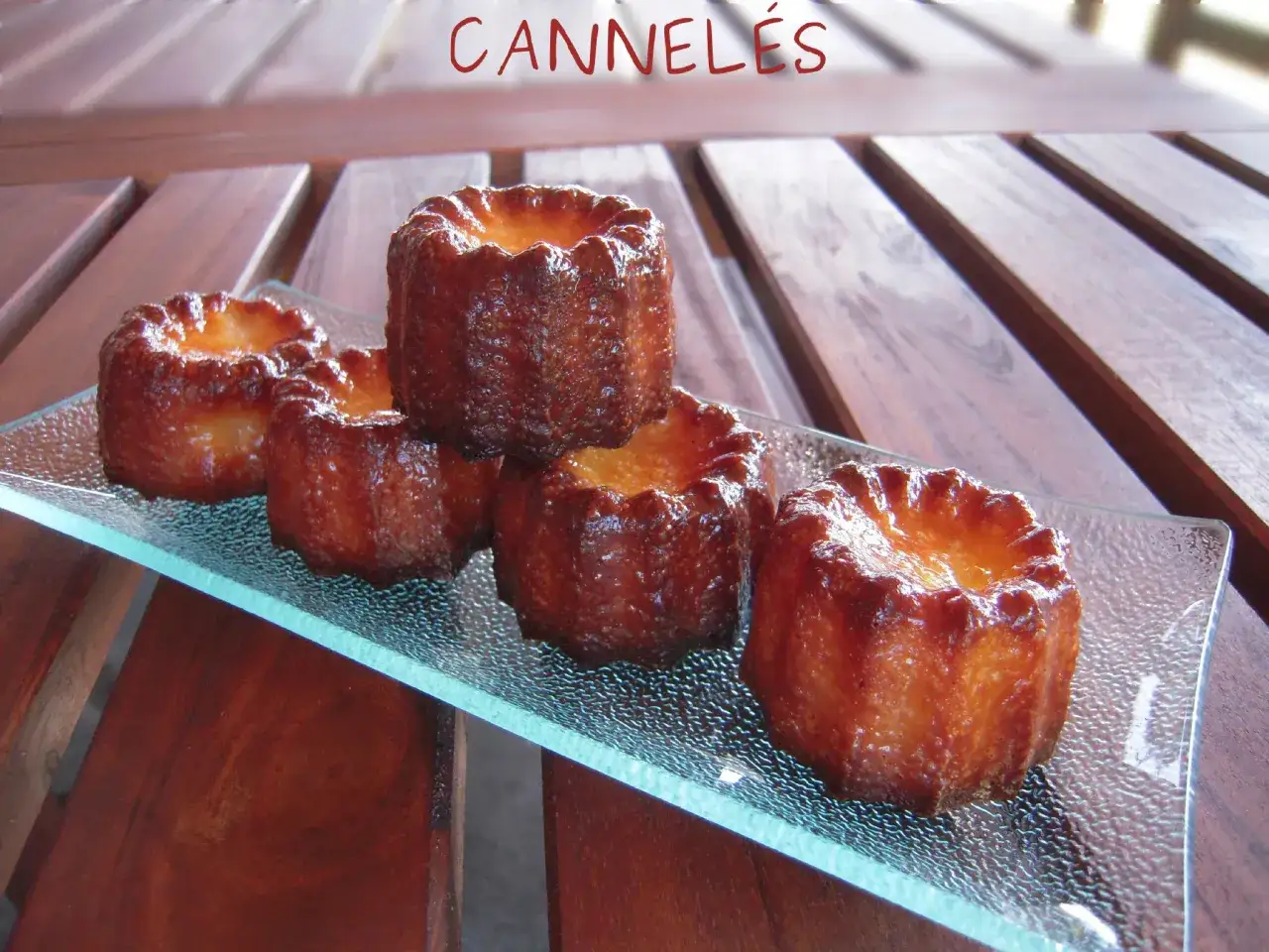Cinq cannel&eacute;s dor&eacute;s, parfaits pour une recette mini cannel&eacute;s moule silicone. Ils sont pr&eacute;sent&eacute;s sur un plateau en verre textur&eacute;.
