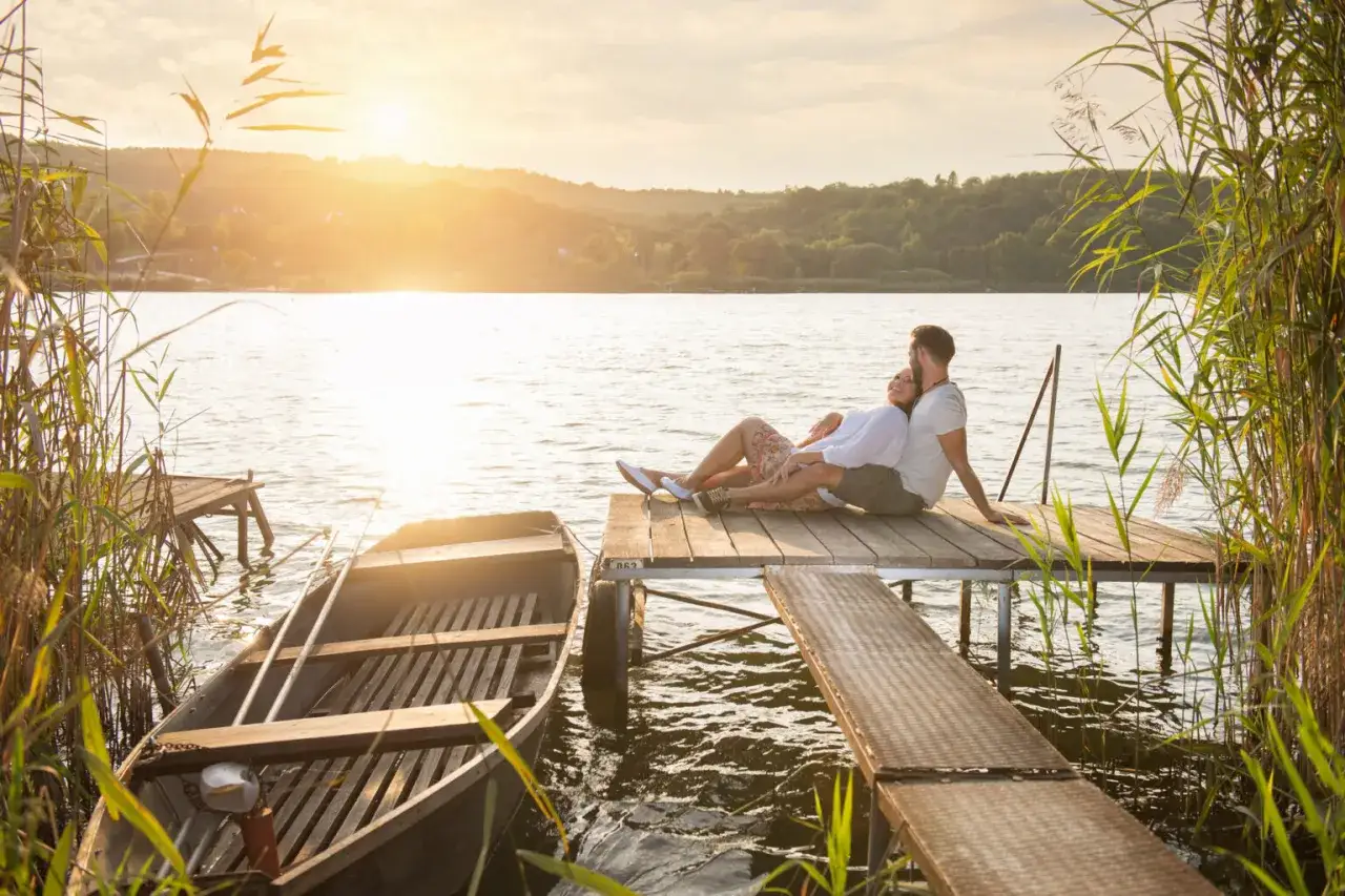 Para, gdzie na Mazury we dwoje? Romantyczny zach&oacute;d słońca nad jeziorem, para siedząca na pomoście, ł&oacute;dka przy brzegu.