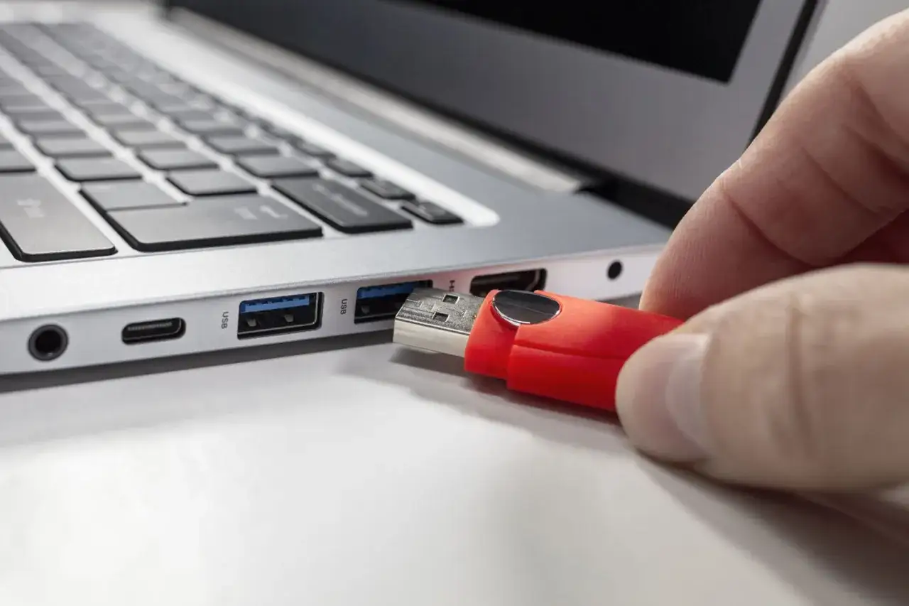 Dłoń wkłada czerwony pendrive do portu USB laptopa. Prosty sposób, jak znaleźć pendrive na laptopie.
