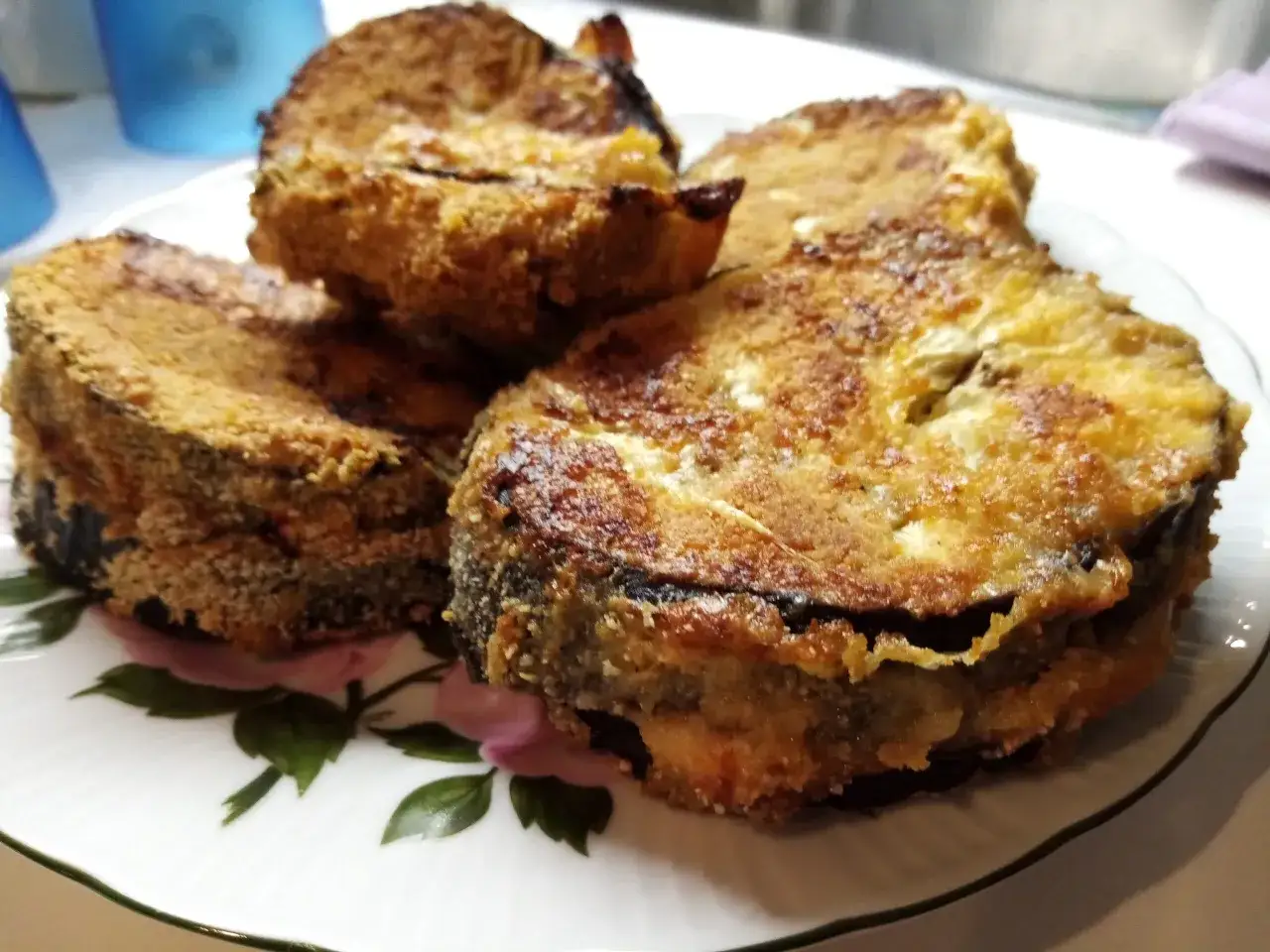 Quattro cordon bleu di melanzane dorati e croccanti, impilati su un piatto bianco con decorazioni floreali.