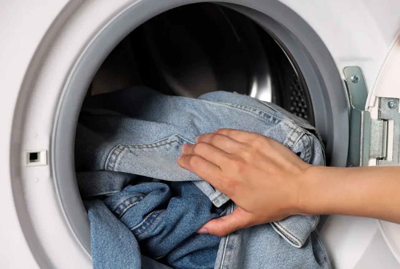 Ręka wkłada dżinsy do pralki. Program denim w pralce zadba o Twoje ulubione spodnie.
