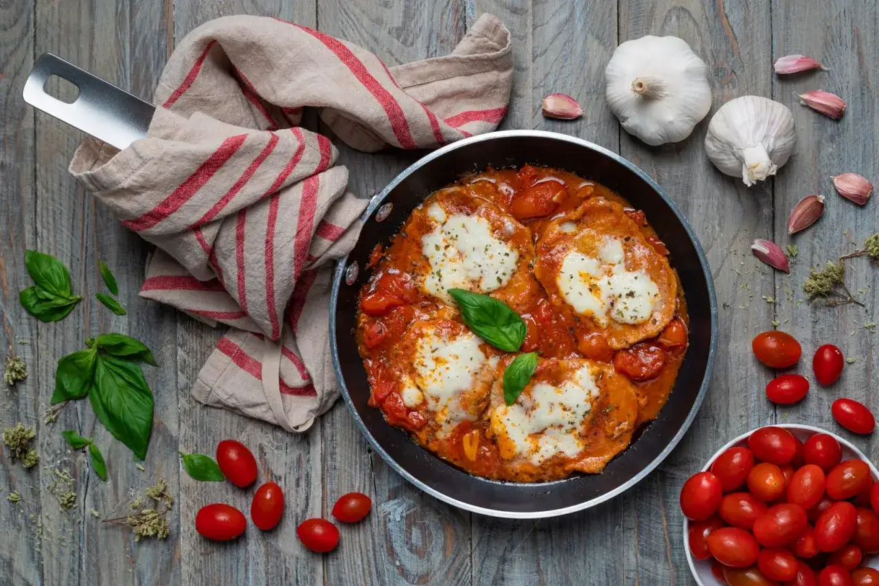 Pizzaiola con mozzarella in padella, contornata da pomodorini, basilico e aglio su un tavolo di legno.