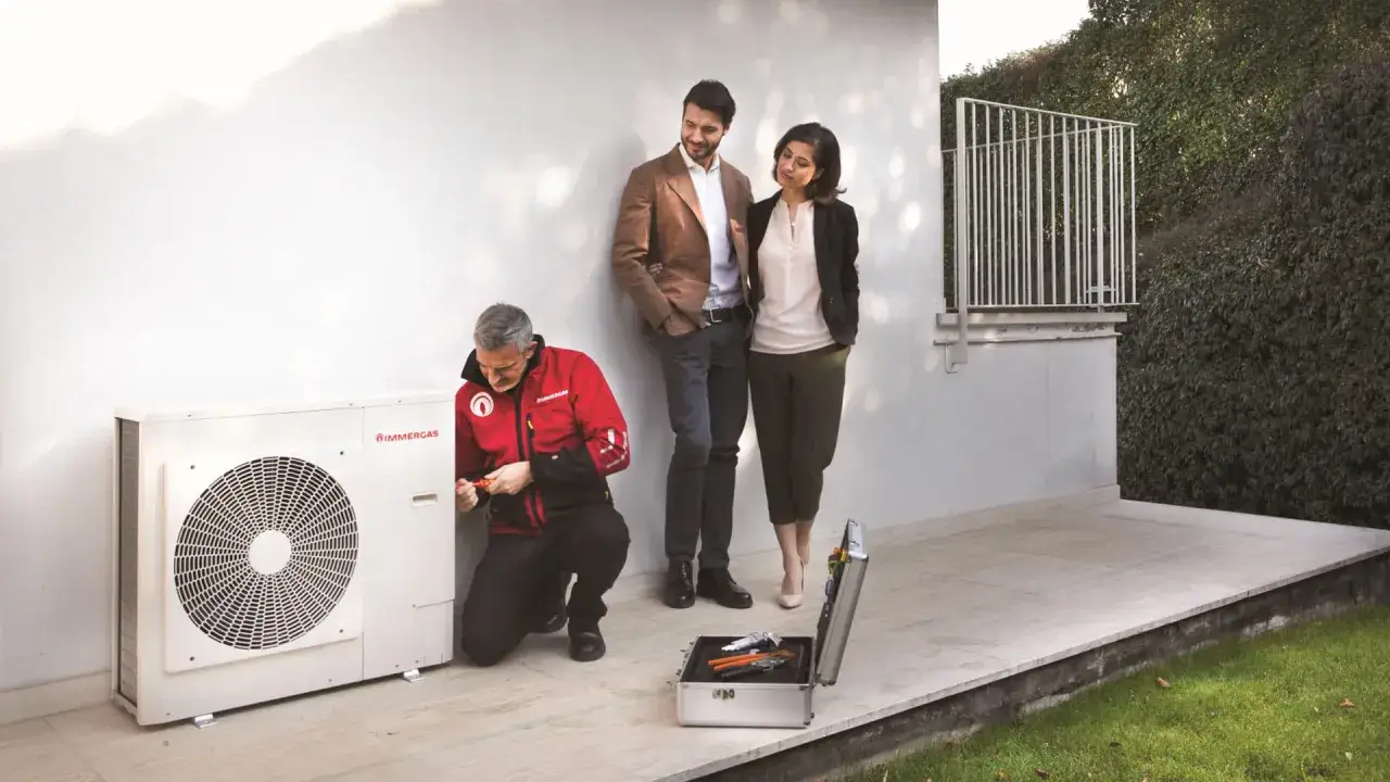 Instalator w czerwonej kurtce naprawia pompę ciepła, podczas gdy para obserwuje.