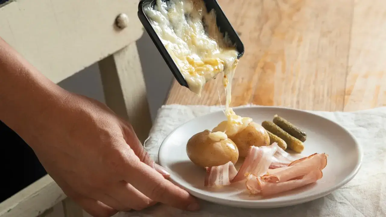 Roztopiony ser z raclette spływa na ziemniaki, obok plastry wędliny i ogórki.