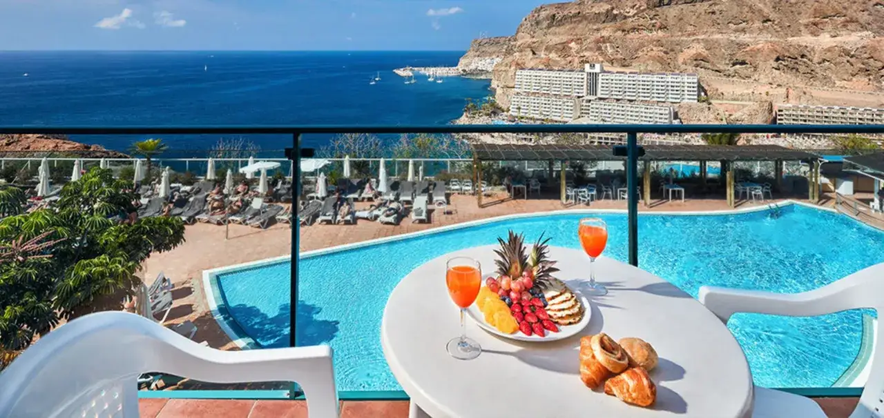 Genießen Sie den Ausblick vom Riu Palace Meloneras: Pool, Meer und ein Frühstückstablett mit Früchten und Gebäck.