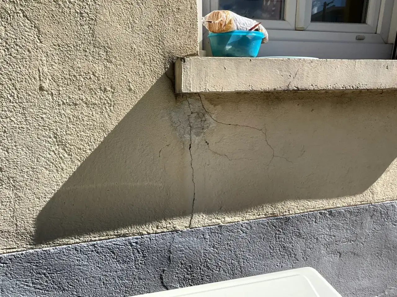 Fissures maison : une ombre triangulaire souligne des fissures sur le crépi d'une maison, près d'un rebord de fenêtre avec un bol bleu.