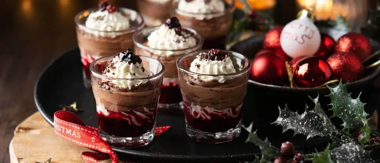 Schokoladenmousse-Desserts in Gläsern, garniert mit Sahne und Beeren, als perfekter weihnachts nachtisch. Weihnachtliche Dekorationen im Hintergrund.