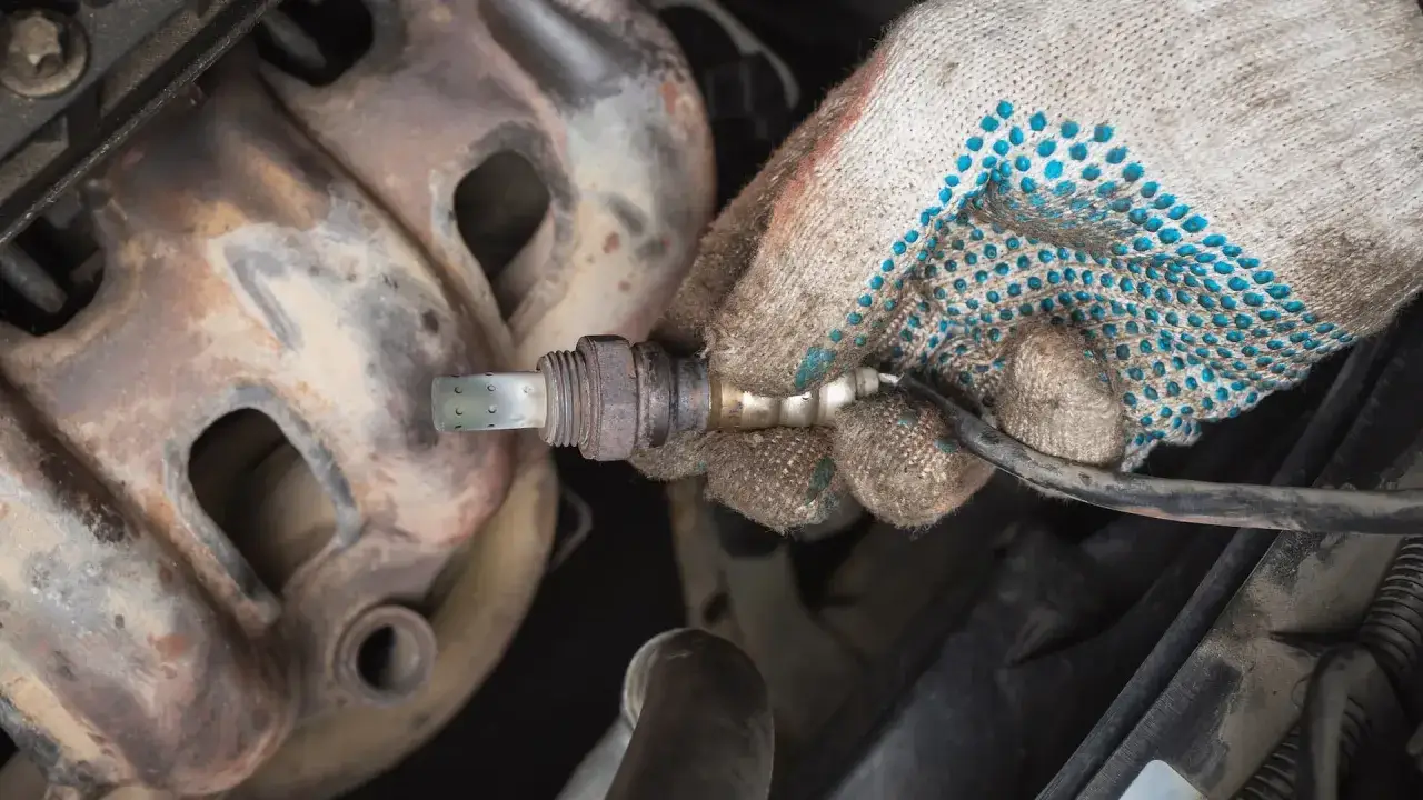 Mechanic's gloved hand holds an oxygen sensor, possibly related to a p0137 code, near a rusty exhaust manifold.