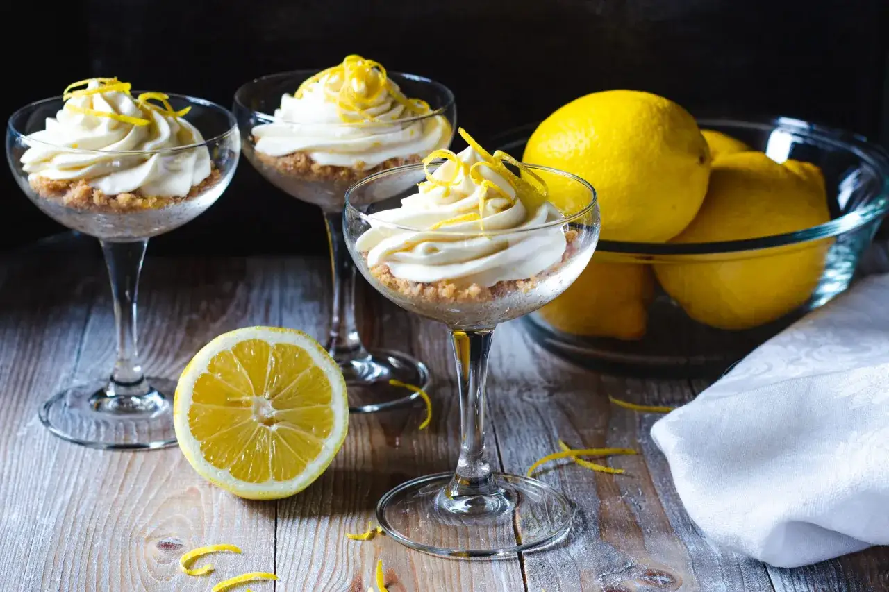 Tre eleganta glas med kr&auml;mig lemon curd, toppad med citronskal. En sk&aring;l med f&auml;rska citroner och en halv citron p&aring; bordet.