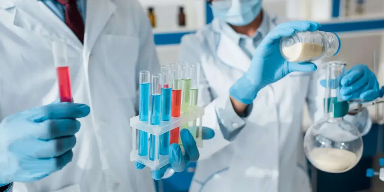 Diagnosta laboratoryjny w niebieskich rękawiczkach przygotowuje próbki do analizy. W probówkach kolorowe płyny, a w kolbie proszek.