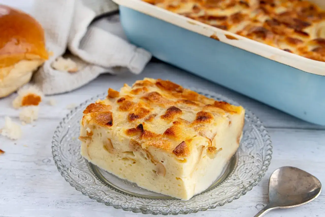 Ein St&uuml;ck goldbrauner Bread Pudding auf einem Glas Teller, daneben ein Laib Brot und eine Auflaufform.
