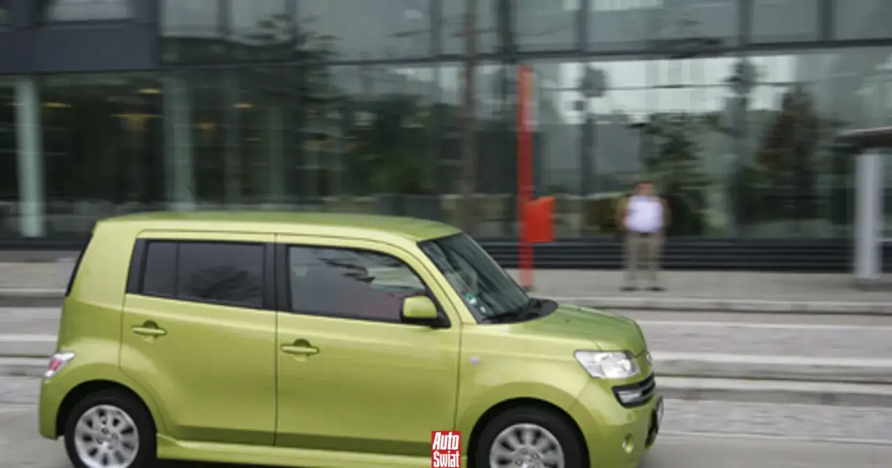 Zielony Daihatsu porusza się po ulicy. W tle nowoczesny budynek i stojący mężczyzna.