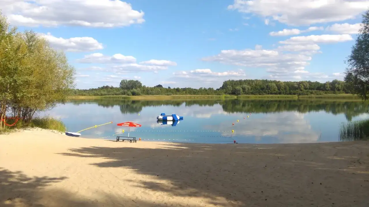 Piękny dzień nad jeziorem Wilczyńskim. Piaszczysta plaża, spokojna woda, dmuchana wyspa i czerwony parasol zapraszają do wypoczynku.