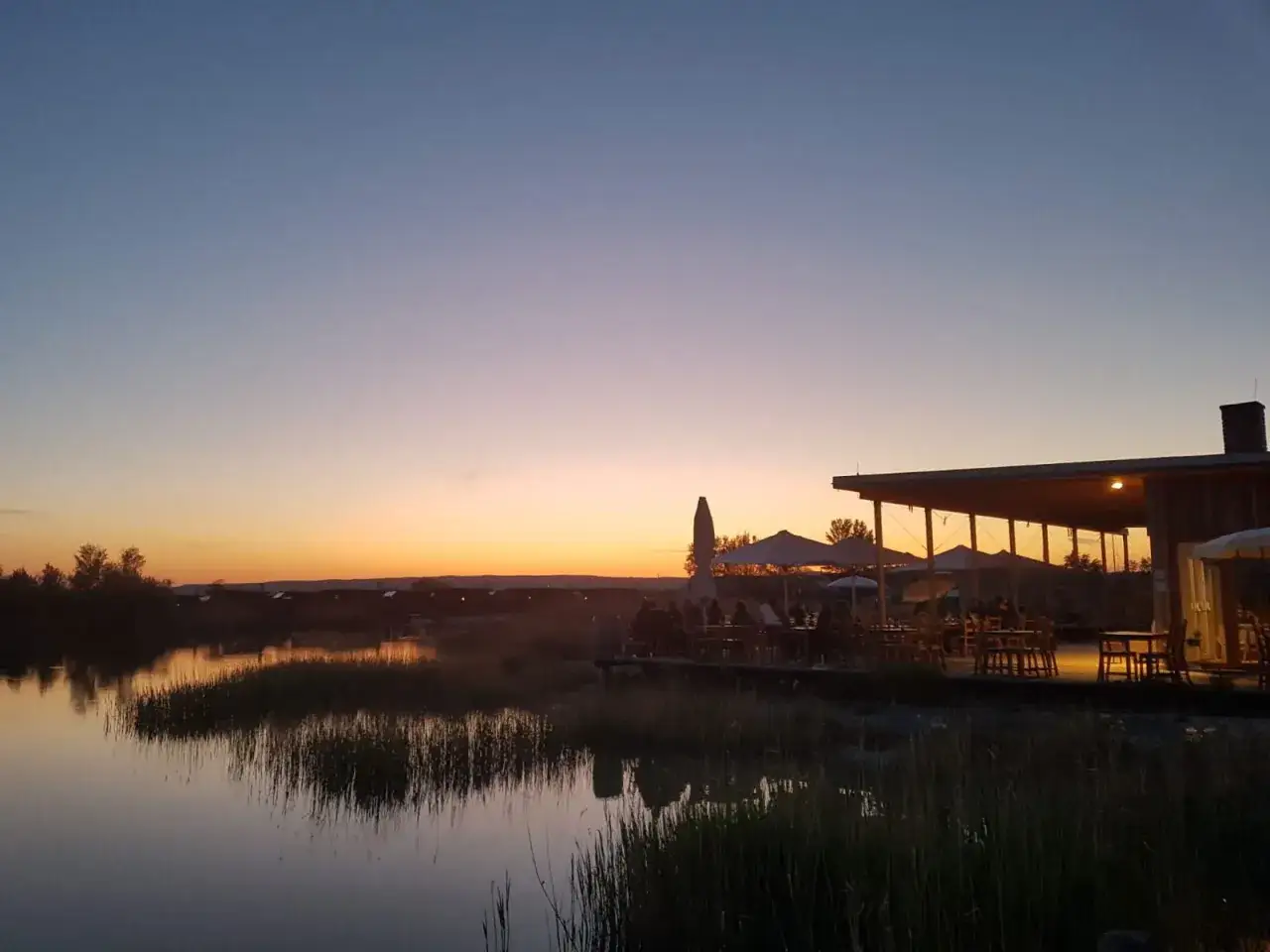 Abendstimmung an einem Restaurant am See. Der Himmel färbt sich in warmen Tönen, während Gäste unter Sonnenschirmen sitzen.