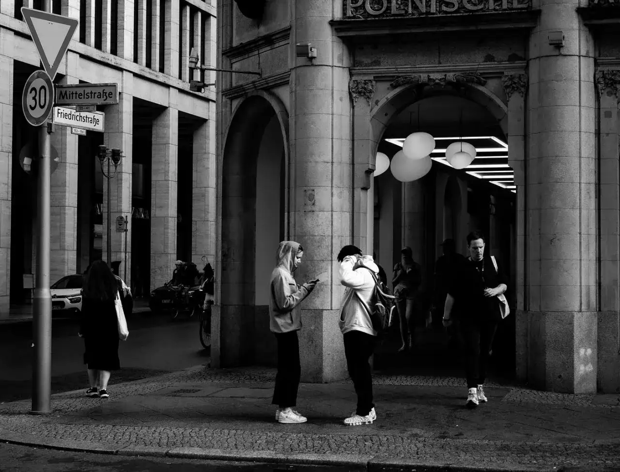 Street photo: Ludzie na chodniku przy ulicy Mittelstraße, w tle łuki budynku i znaki drogowe.