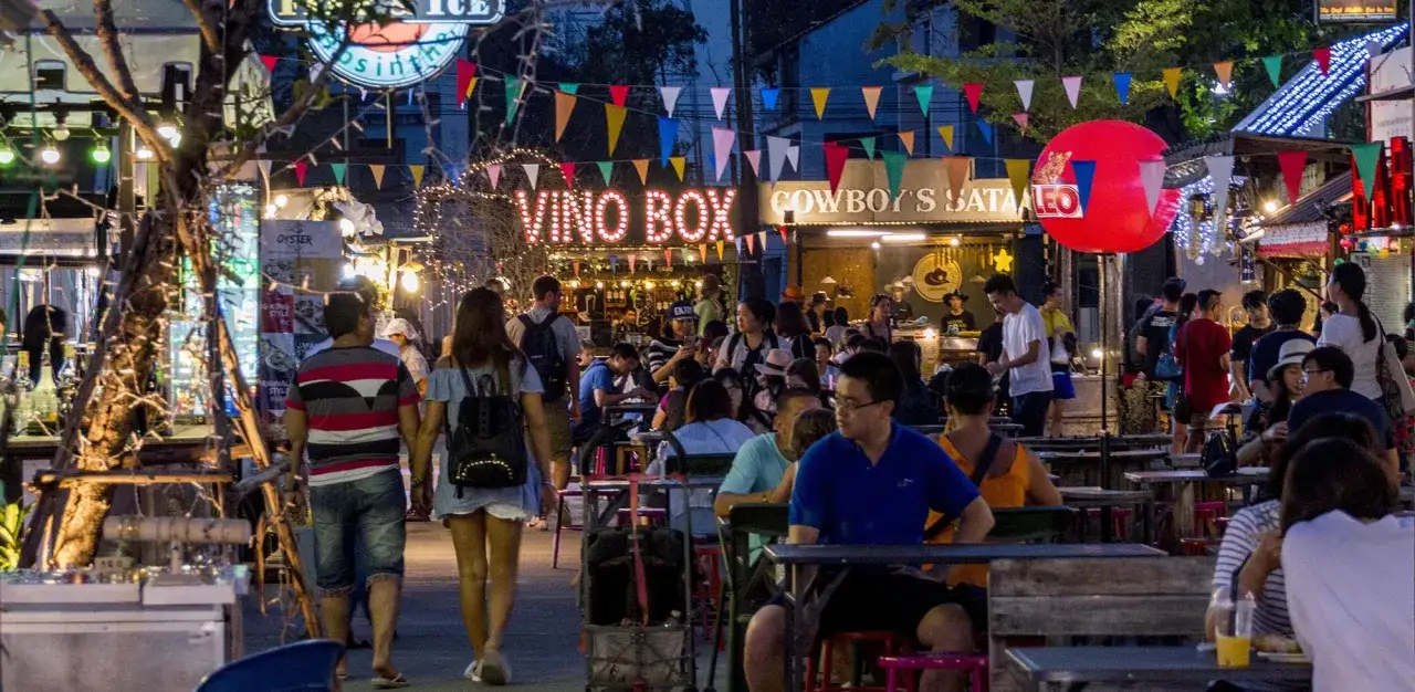 Tętniący życiem nocny targ w Chiang Mai. Ludzie siedzą przy stolikach, jedzą i piją pod kolorowymi flagami i neonami.