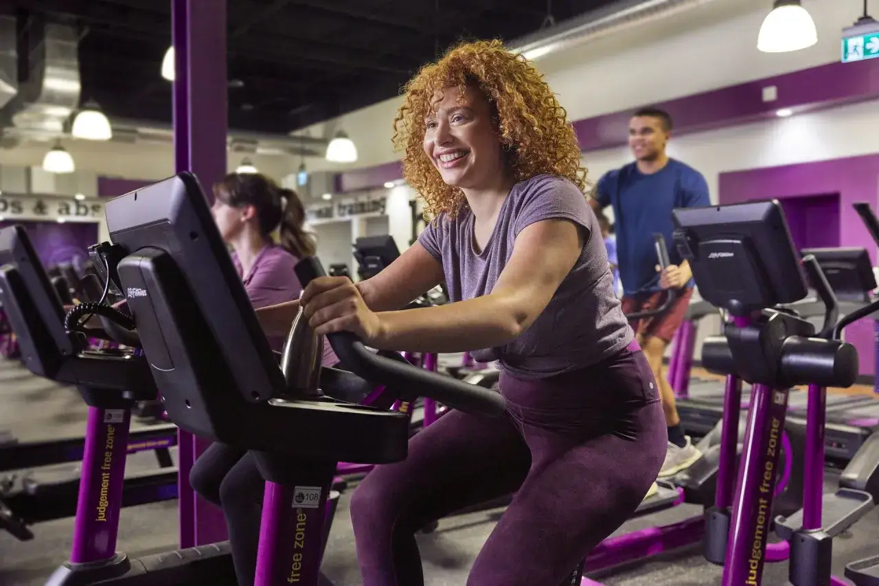 Uśmiechnięta kobieta z kręconymi włosami wykonuje trening cardio na rowerku stacjonarnym na siłowni.