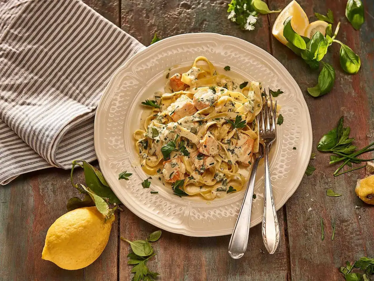 Cremige Pasta al Salmone mit Lachs, Kräutern und Zitrone auf einem Teller, daneben eine Serviette und frische Zitronenspalten.