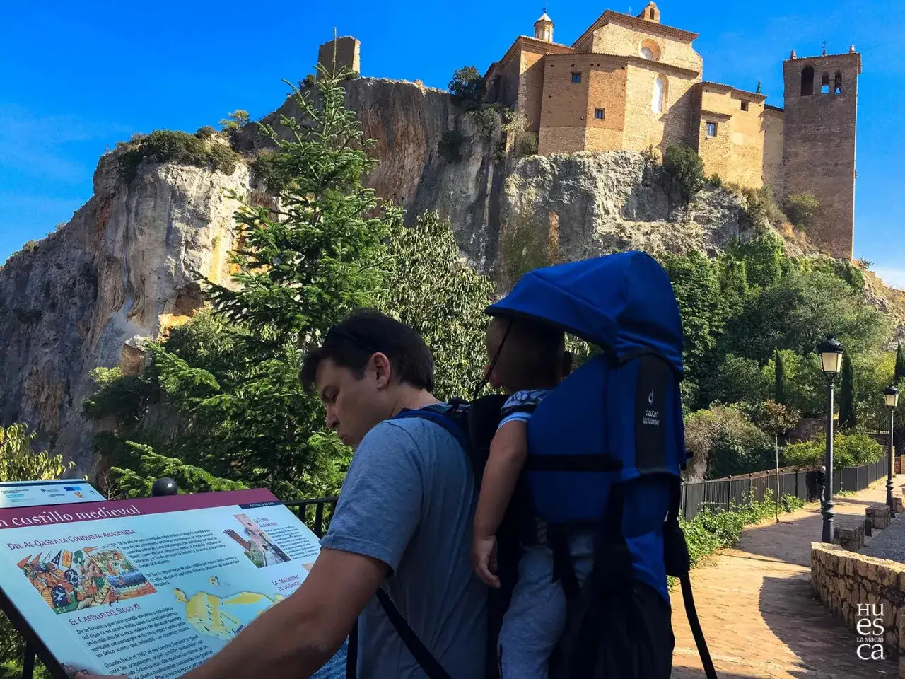 Padre con beb&eacute; en mochila leyendo un panel informativo sobre un castillo medieval en Huesca con ni&ntilde;os.