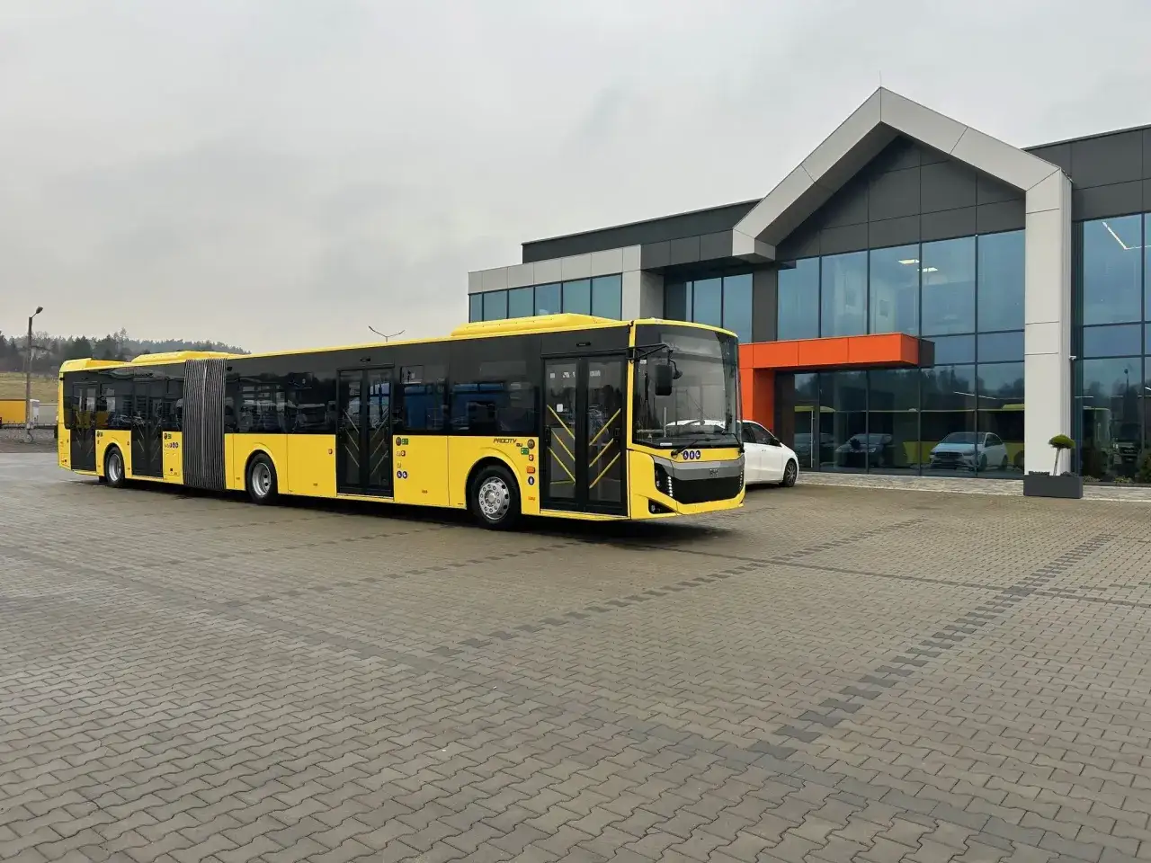 Ż&oacute;łty przegubowy autobus miejski przed nowoczesnym budynkiem. Trudno określić, ile bus ma miejsc, ale jest bardzo długi.