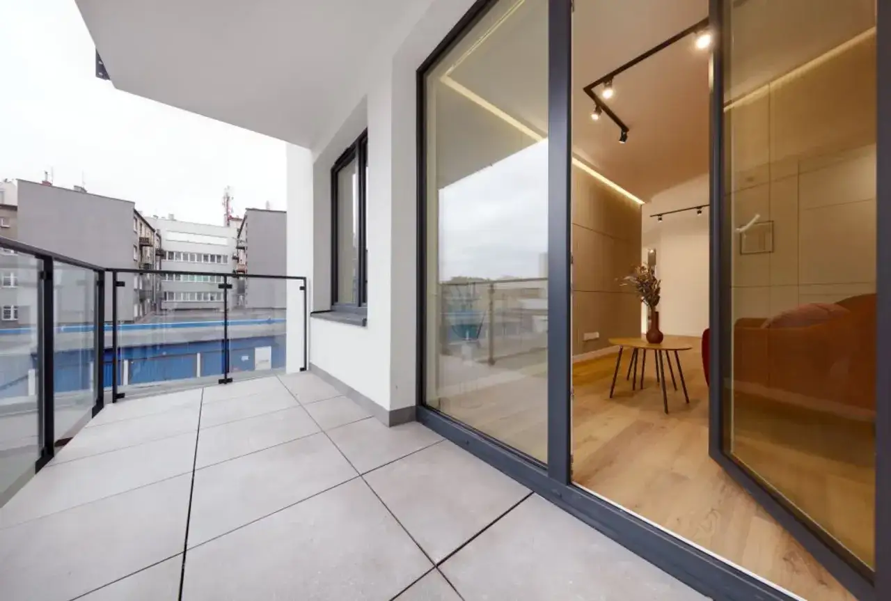 Nowy balkon z płytkami i szklaną balustradą. Wnętrze z drewnianą podłogą i kanapą. Idealne miejsce na chwilę relaksu i ucieczkę od miejskiego zgiełku.