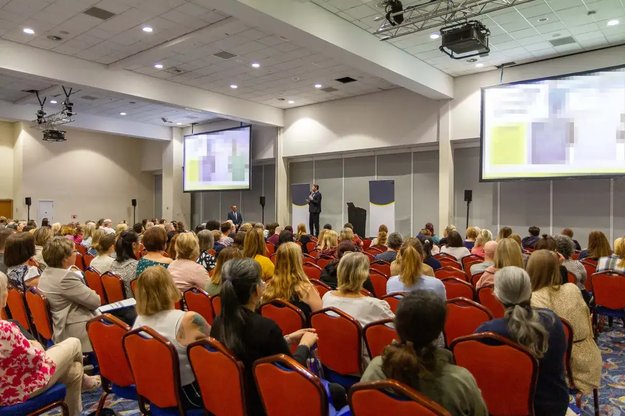 Conference photography captures a speaker addressing a seated audience in a large hall.
