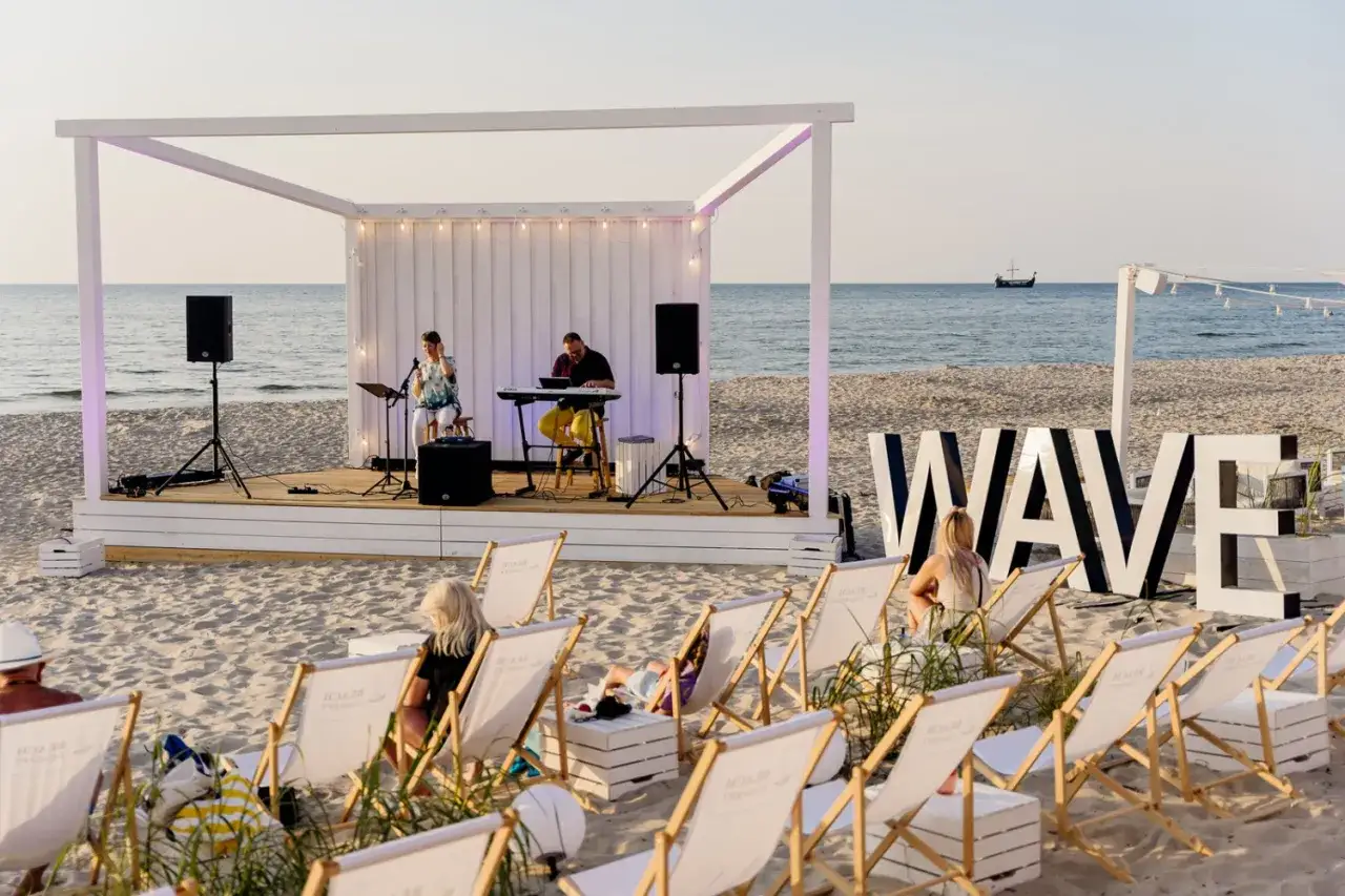 Koncert na plaży w Międzyzdrojach. Muzycy grają na scenie, a widzowie siedzą na leżakach. Wielkie litery