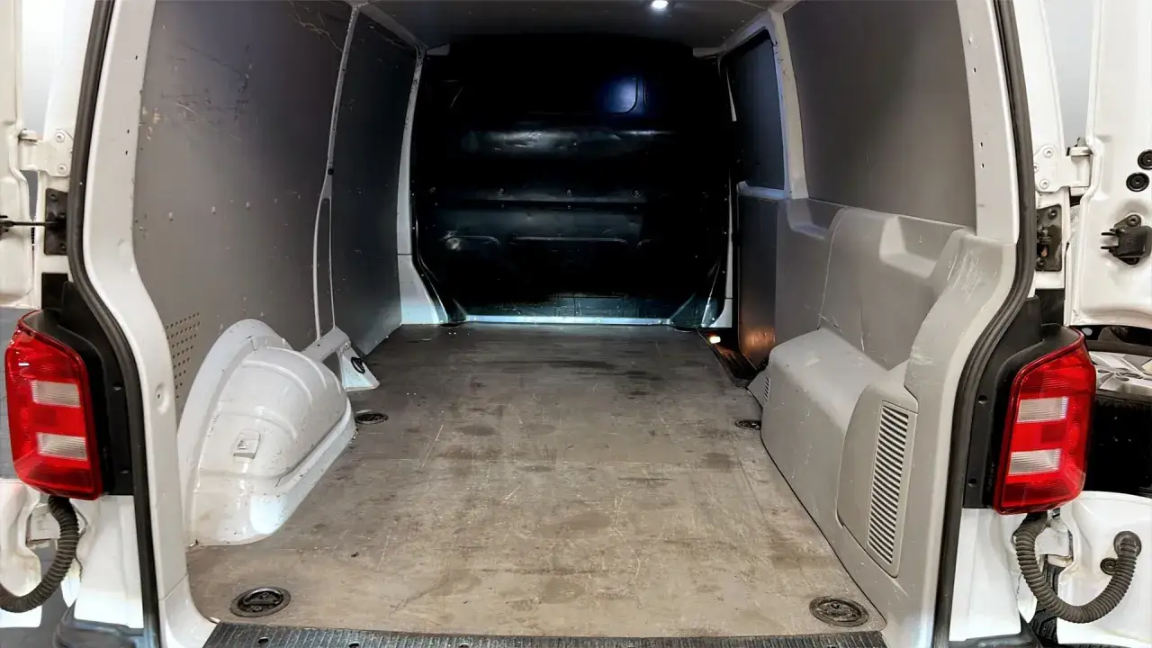 Insidan av en vit, begagnad Volkswagen Caravelle. Lastutrymmet &auml;r tomt med gr&aring; v&auml;ggar och ett slitet tr&auml;golv.