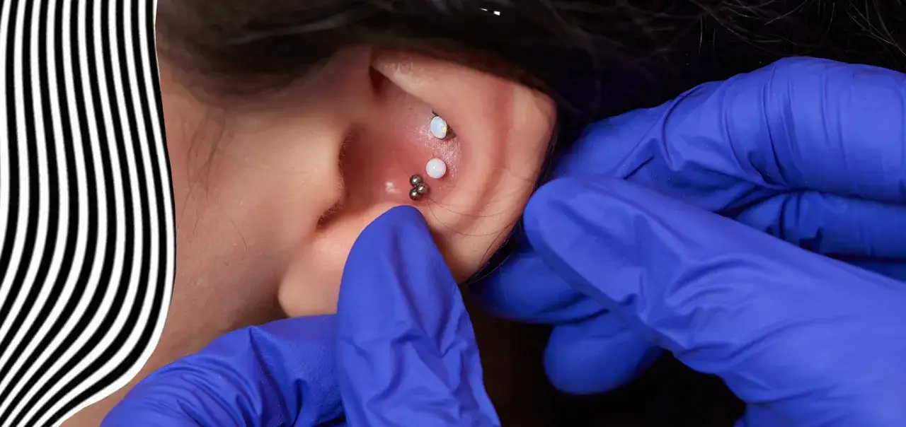 N&auml;rbild p&aring; ett &ouml;ra d&auml;r en piercare med bl&aring; handskar s&auml;tter in smycken. En opal och en klunga av sm&aring; kulor pryder &ouml;rat, en modern achilles piercing.