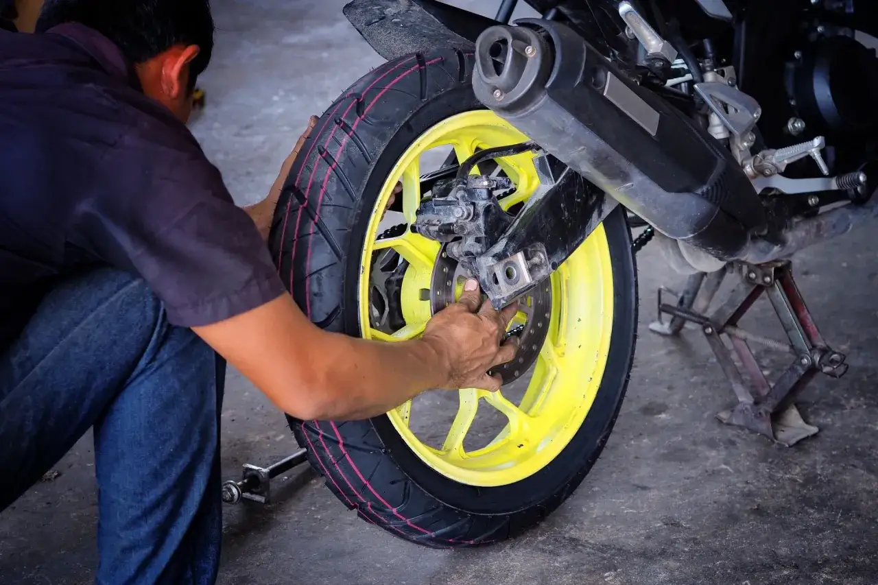 Mechanik dokonuje wymiany opony motocyklowej, pracując przy ż&oacute;łtej feldze i hamulcu.