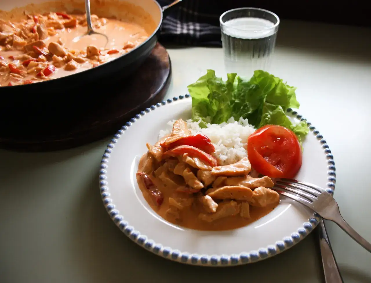 En tallrik med kyckling stroganoff, ris, sallad och tomat. En kastrull med mer stroganoff syns i bakgrunden.