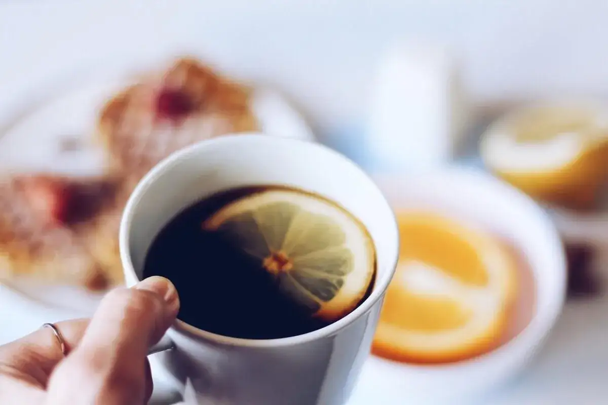 Eine Hand hält eine Tasse mit schwarzem Kaffee mit Zitrone. Im Hintergrund sind Pfannkuchen und Orangenscheiben zu sehen.