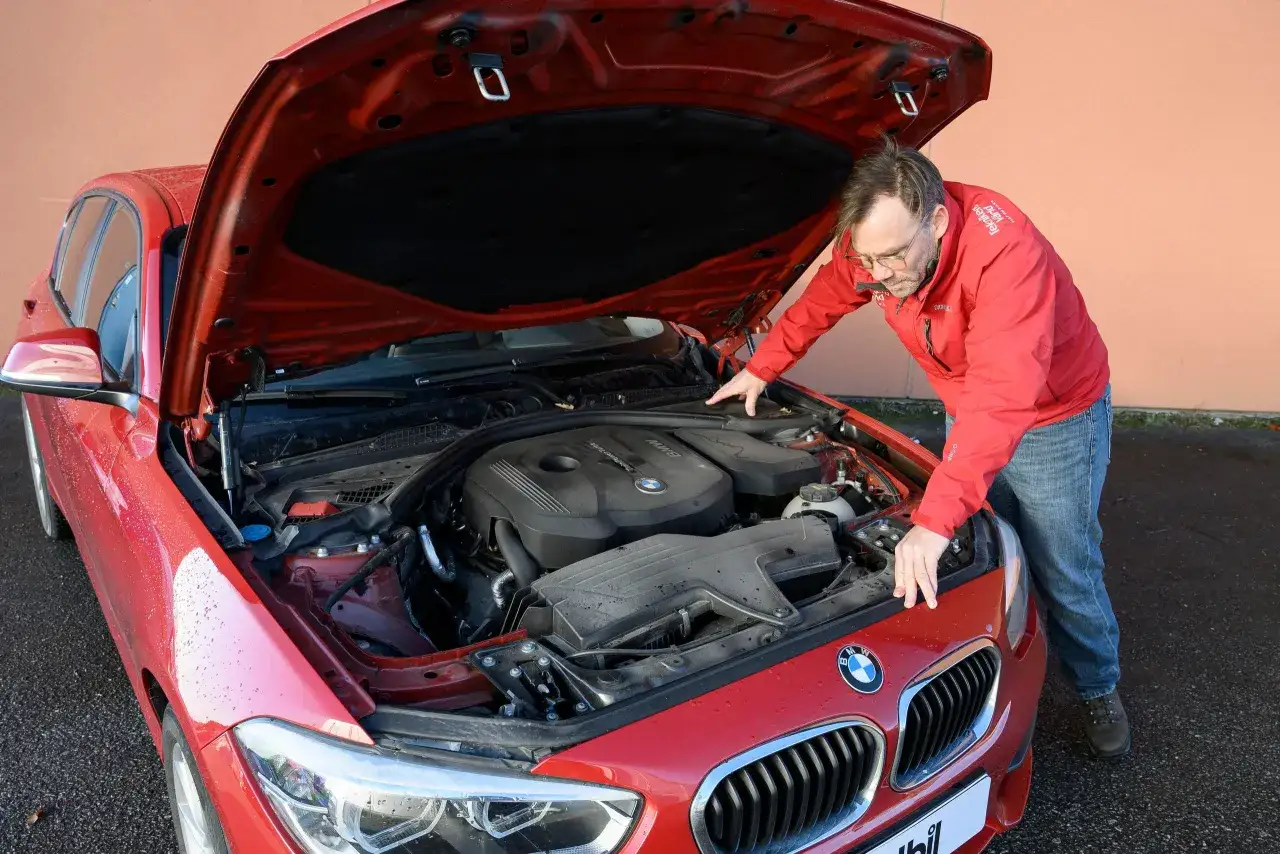 Man inspekterar motorn på en röd, begagnad BMW 1-serie. Motorhuven är öppen.