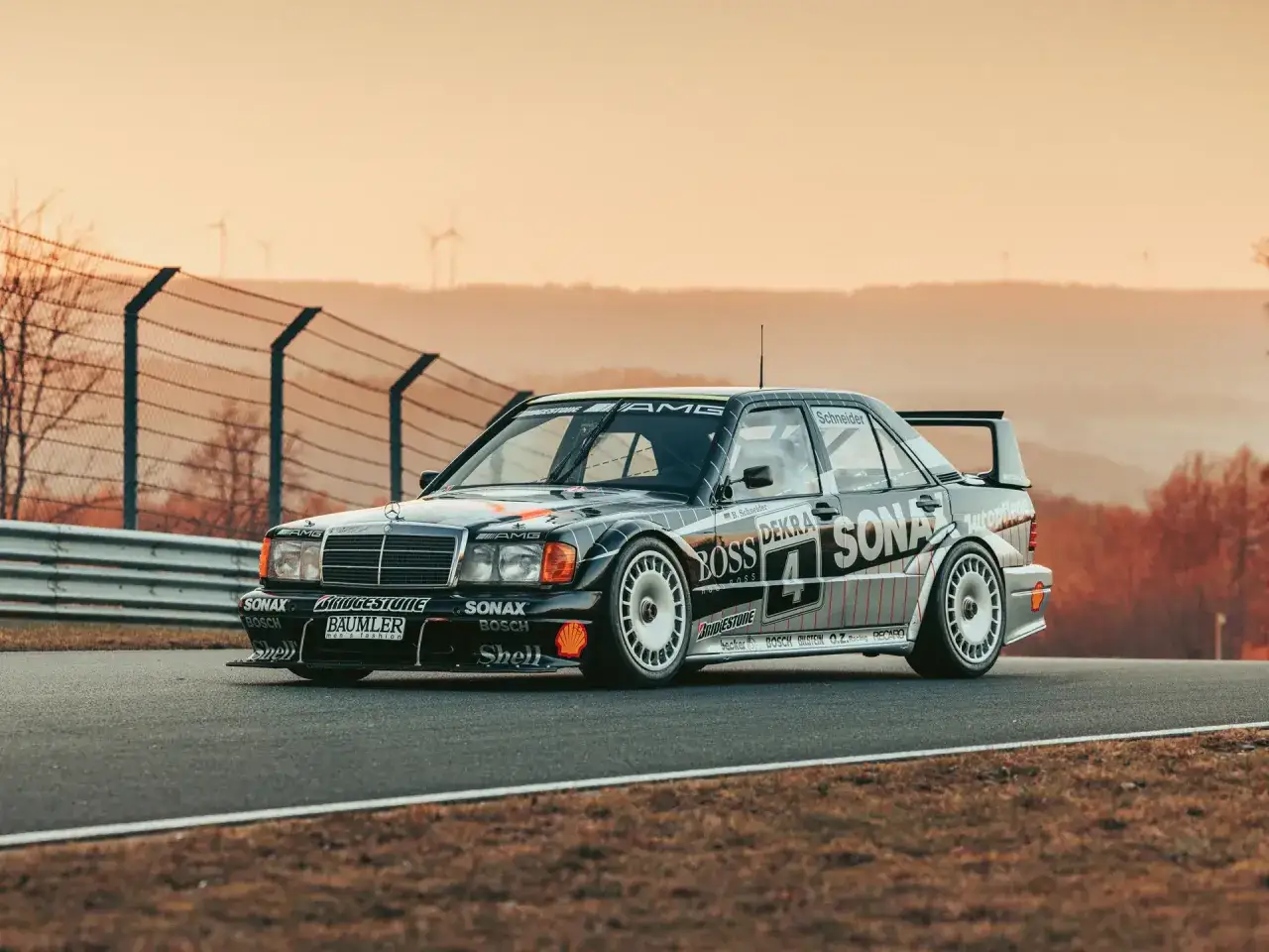 Klasyczny Mercedes 190e Evo 2 na torze wyścigowym, z logotypami sponsorów i w tle wiatrakami.