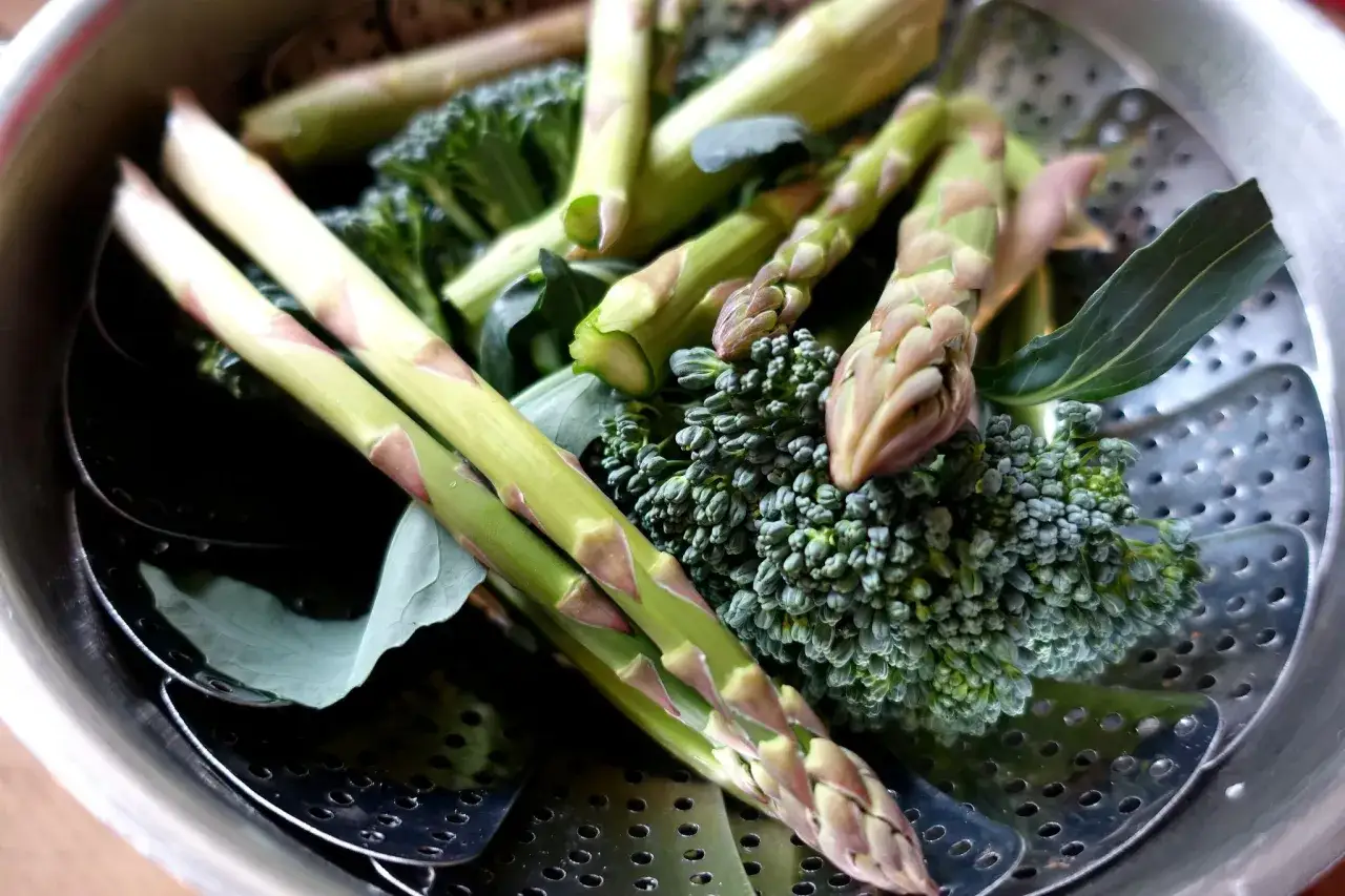 Färska sparrisar och broccoli ligger i en ångkokare, redo för ett hälsosamt ångkokare recept.