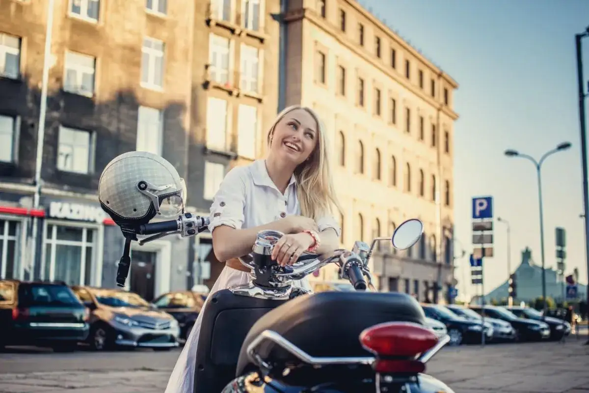 Uśmiechnięta blondynka na skuterze diesel, z hełmem na kierownicy, na tle kamienic i samochodów.