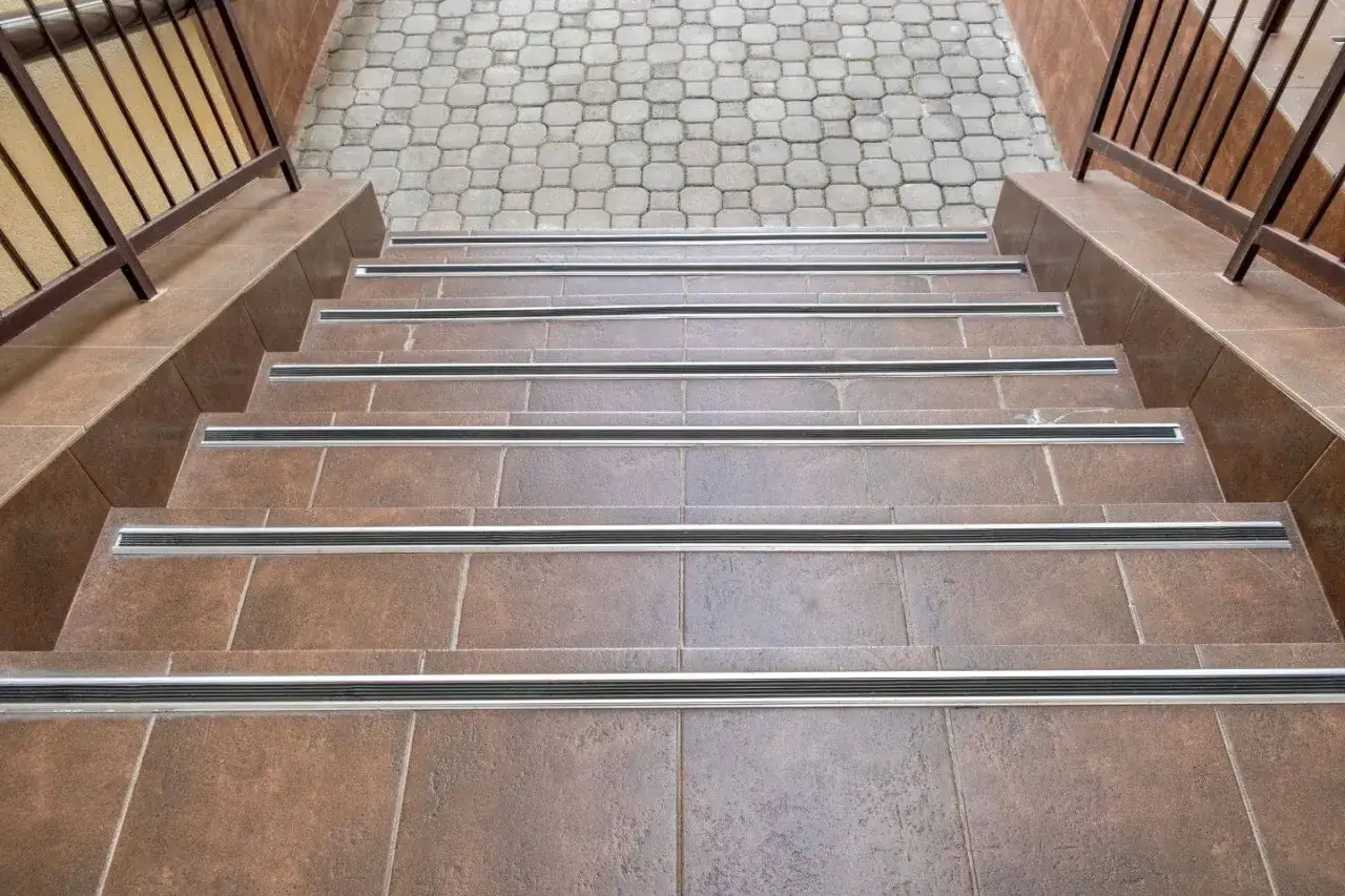 Schody z płytek ceramicznych w brązowym kolorze, z metalowymi antypoślizgowymi listwami na każdym stopniu.