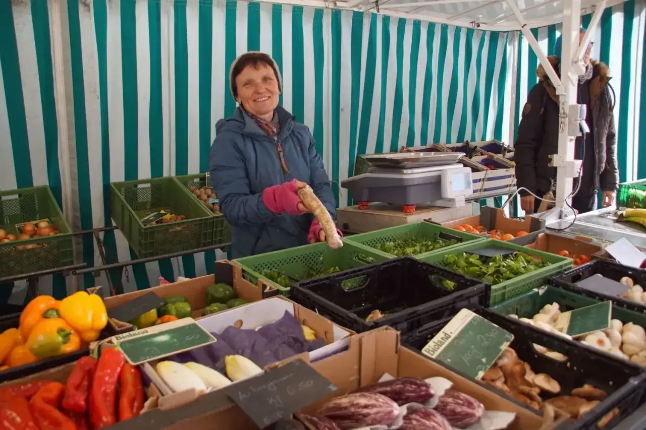 Verkäuferin hält Meerrettich, der sich vom Rettich unterscheidet. Auf dem Markt gibt es auch Paprika, Chicorée und Knoblauch.