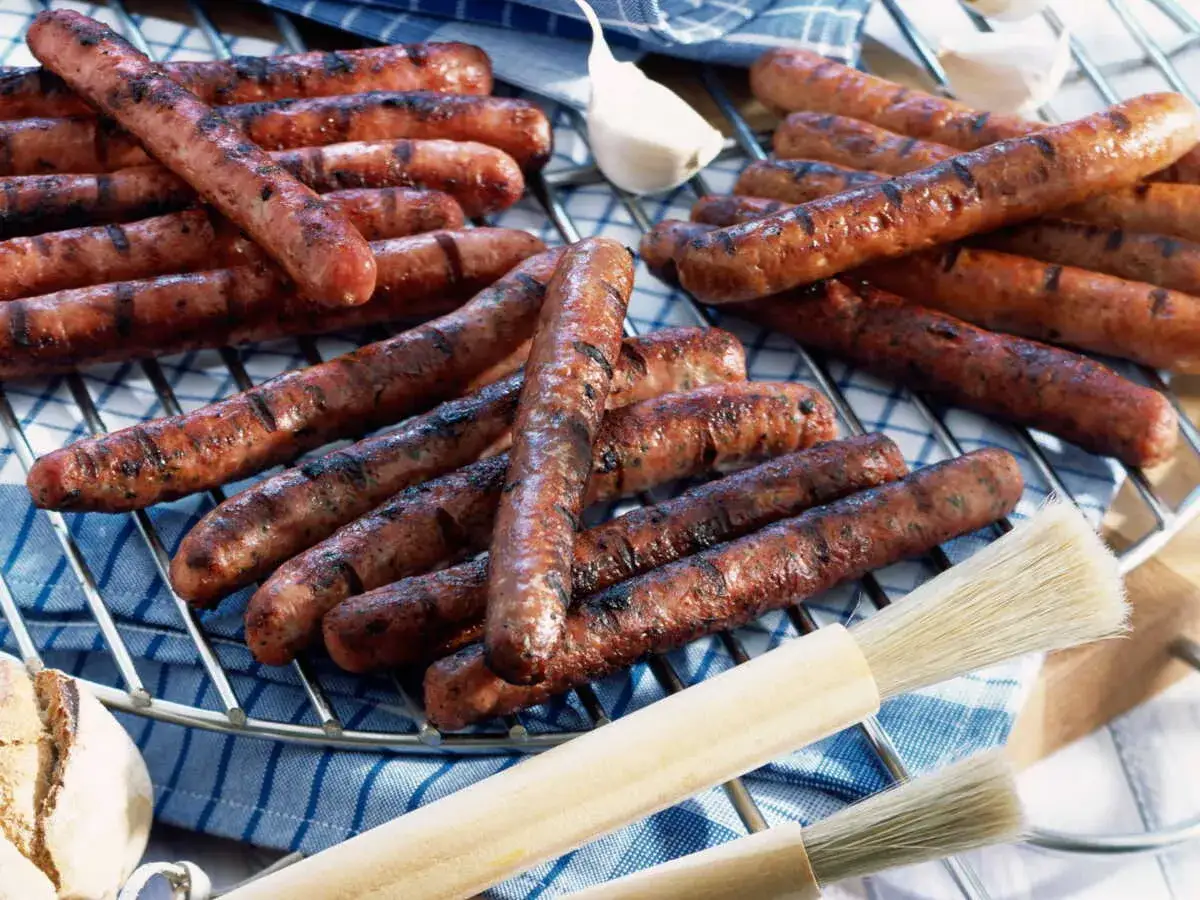 Merguez grillées sur un barbecue, prêtes pour le temps de cuisson. Des brosses de cuisine et de l'ail sont à proximité.