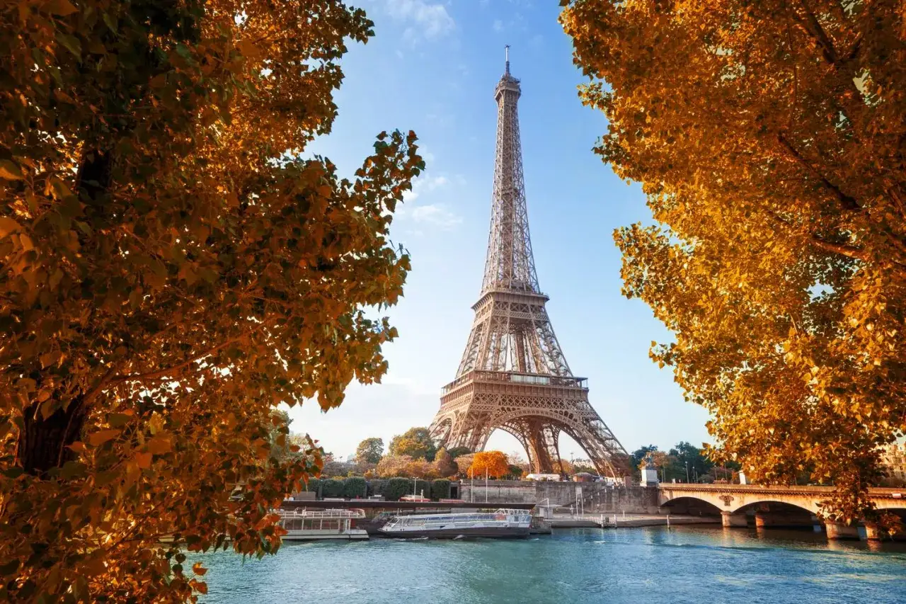 Eiffelturm im Herbstlaub, die Seine mit Booten und eine Brücke. Die goldene Färbung der Blätter deutet auf die beste Reisezeit Paris für malerische Ausblicke hin.