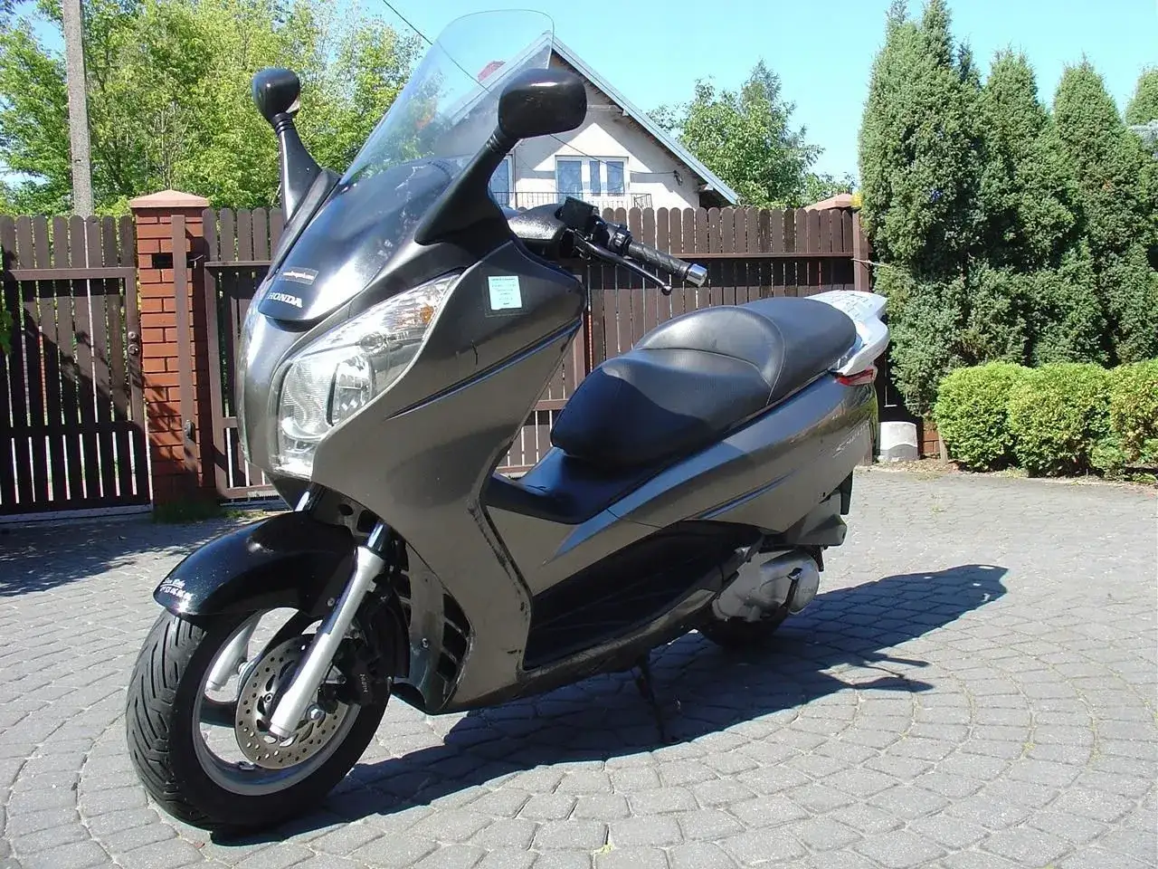 Szary skuter Honda Swing 125 zaparkowany na kostce brukowej, z drewnianym płotem i zielenią w tle.