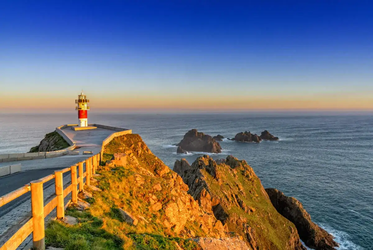 Faro rojo y blanco en un acantilado rocoso, con el mar azul y el cielo al atardecer. Un camino serpentea hacia &eacute;l, ideal para una ruta por Galicia.