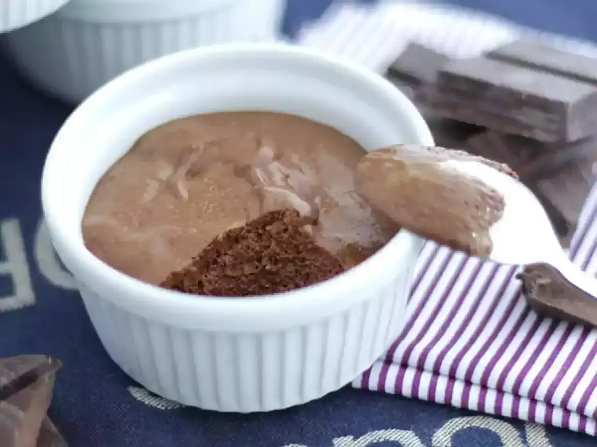 Une cuillère prélève une portion aérienne de mousse au chocolat dans un ramequin blanc. Des morceaux de chocolat noir sont disposés à côté.