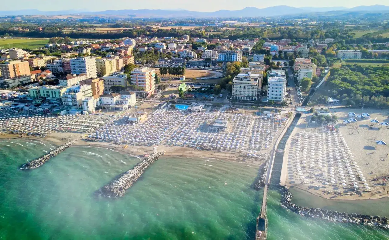 Vista aerea di una spiaggia affollata a Cattolica, con file ordinate di ombrelloni bianchi e blu. Il mare turchese lambisce la costa, mentre sullo sfondo si intravedono le colline.